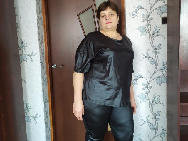 ElzaJewel with short brown hair, black shiny t-shirt, and black leather pants, standing in front of a wooden door, looking straight at the camera with a neutral expression.