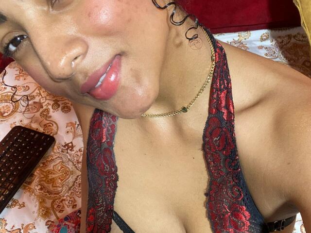 NatalyBlonder with curly black hair, tan skin, pink lips, and a gold necklace, wearing a red and black lace bra, showing her cleavage and a subtle smile.