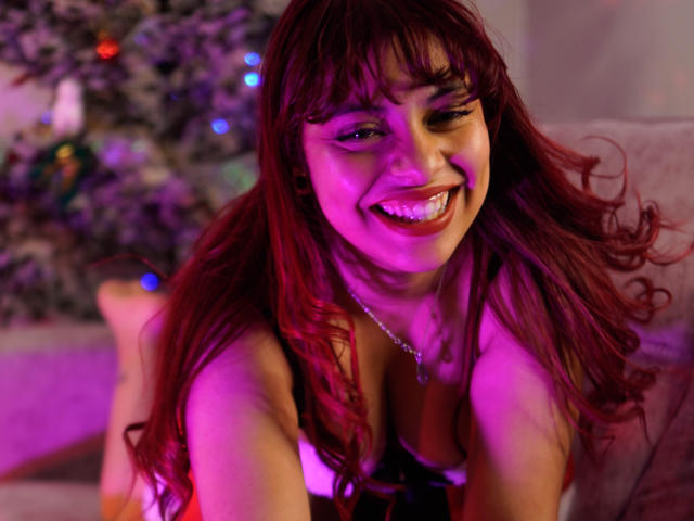 Agathadebella with red hair, smiling, winking, wearing a low-cut top, showing her tits, pink and purple light, necklace, sitting on a couch, Christmas tree in the background.