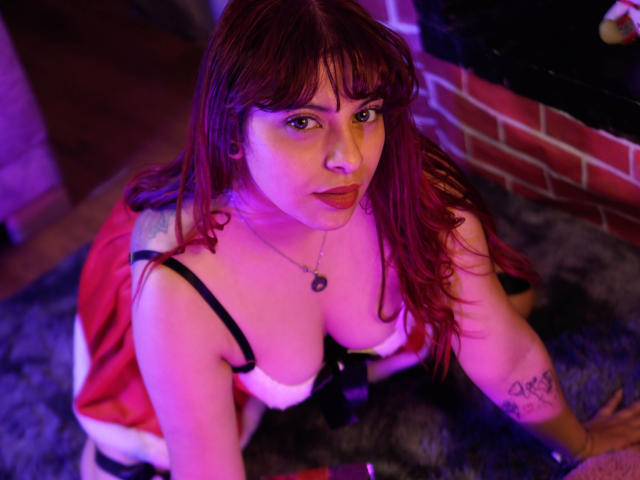 Agathadebella with red hair, large boobs, and a tattoo on her arm, wearing a Santa outfit, kneeling on a purple carpet, looking up with pink and purple lighting.