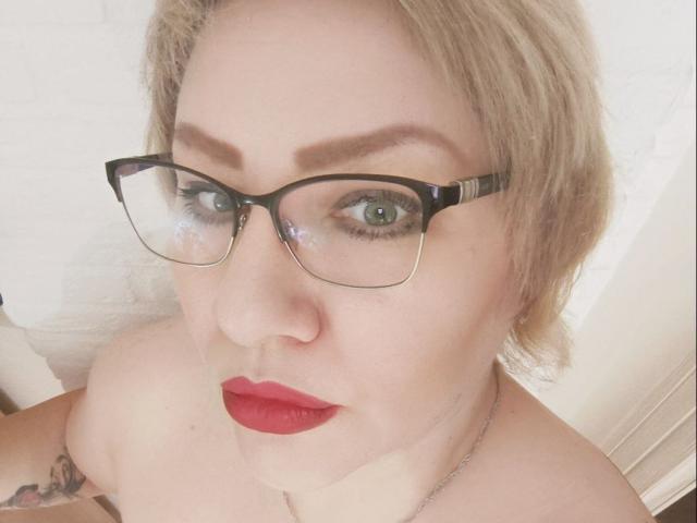 AngelinaGlam with blonde hair, glasses, red lipstick, green eyes, and a tattoo on her left shoulder, looking up at the camera. Boobs visible, slightly blurred.