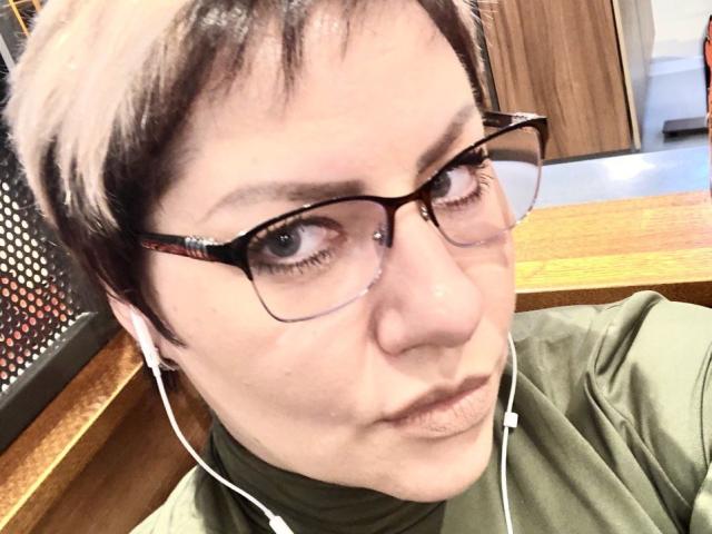 AngelinaGlam with short brown hair, glasses, green shirt, earbuds in, slightly pouty expression, wooden table in background.