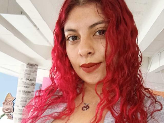 Agathadebella has bright red curly hair, brown eyes, and red lipstick. She's wearing a white top with a pink accent and a necklace with a heart pendant.