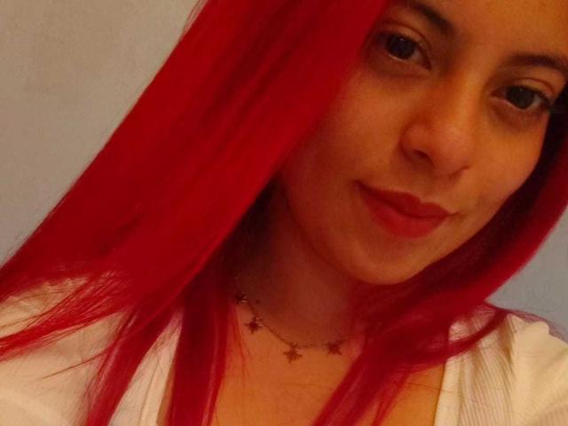 Agathadebella with bright red hair, slight smile, wearing a white top and star necklace, brown eyes, light skin, and subtle makeup. Her tits are small, and she's cute.