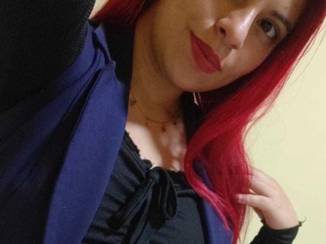 Agathadebella with red hair, wearing a purple cardigan over a black top with a bow, has red lipstick and a necklace, smiling slightly, showing small tits.