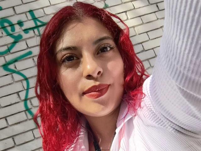 Agathadebella with red curly hair, wearing a white shirt, has a cute face and is smiling with red lipstick. Her arm is raised, taking a selfie.