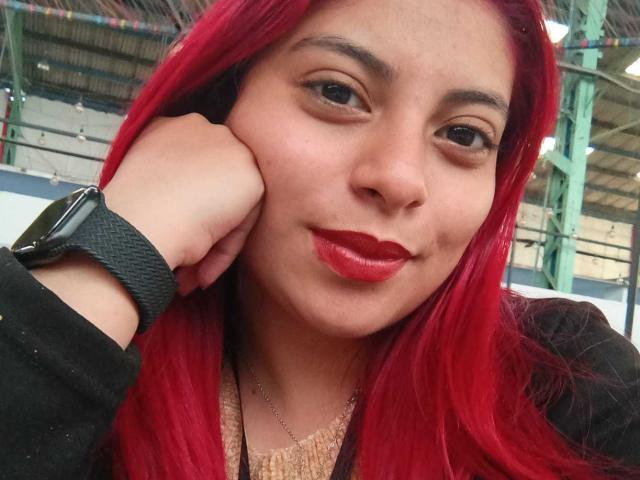 Agathadebella with bright red hair, red lipstick, and black wristwatch, resting her head on her hand, smiling subtly. She's wearing a black top with a gold necklace.