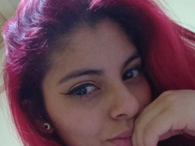 Agathadebella has red hair, blue eyes with black eyeliner, and is biting her finger with a slight smile. She's wearing small gold earrings.