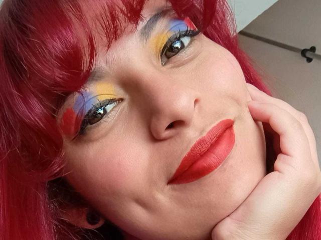 Agathadebella with red hair, yellow-blue-orange eyeshadow, red lipstick, and black ear gauges, smiling with hand near face. Her eyes are reflective, and she's looking at the camera.