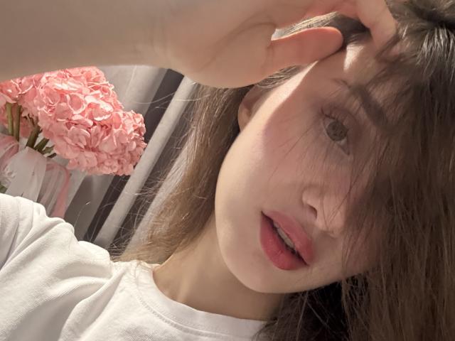 LaraSweetyHot met bruin haar, gedeeltelijk het gezicht bedekkend, wit shirt, roze lippen, hand in haar, roze bloemen op de achtergrond, zacht licht, bleke huid, subtiele make-up, lichte glimlach, ontspannen..