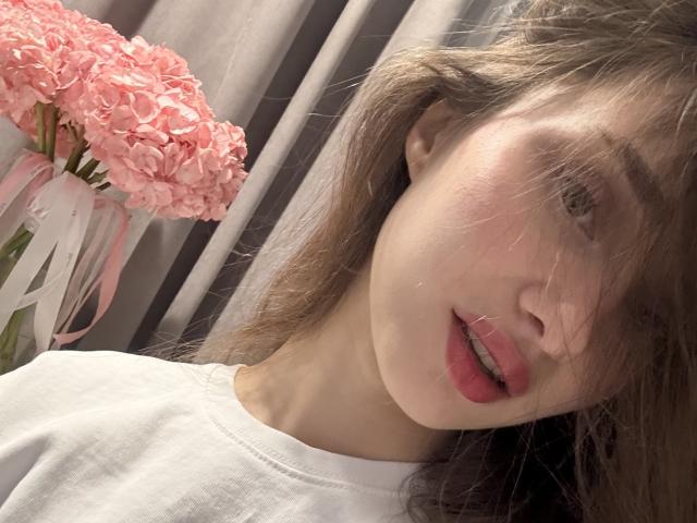Het gezicht van LaraSweetyHot, met roze bloemen op de achtergrond, gekleed in een witte blouse, met bruin haar dat één oog bedekt, lichte huid, roze lippen en een licht geopende mond..