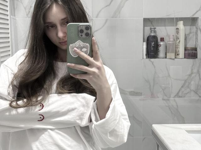 LaraSweetyHot, con cabello castaño largo, vestida con una camisa blanca, tomando una selfie en un espejo en un baño, sosteniendo un teléfono verde a cuadros, con una expresión seria, mostrando ligeramente sus senos..