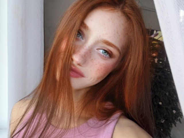LaraSweetyHot a les cheveux roux, les yeux bleus, des taches de rousseur et porte un haut rose, avec un léger sourire et ses cheveux qui couvrent partiellement son visage. Ses seins ne sont pas visibles..