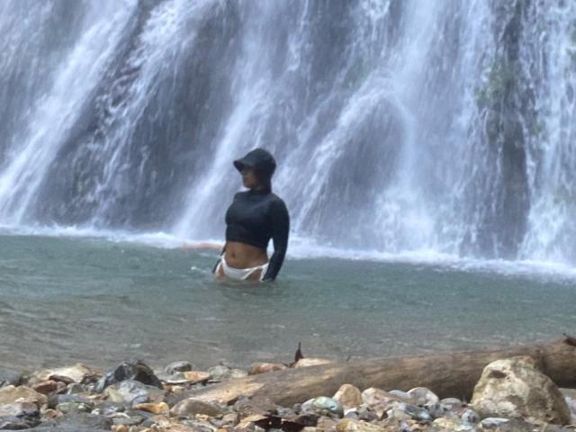AkendaCurls staat tot aan haar middel in een rotsachtige beek, gekleed in een zwart shirt met lange mouwen en witte onderbroek, met een waterval op de achtergrond en een boomstam op de voorgrond..