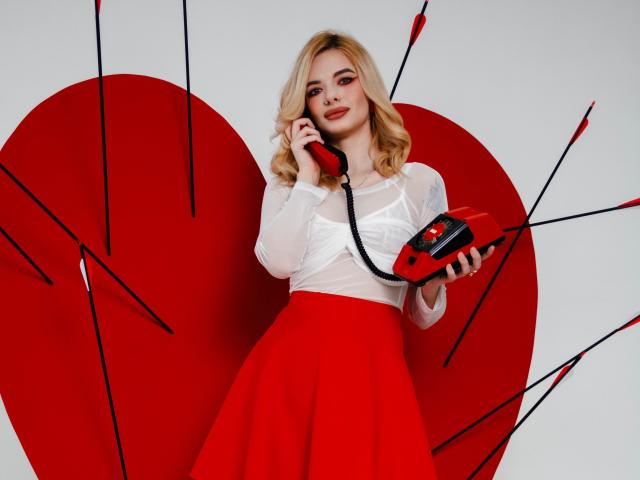 AdeleClifford, con cabello rubio, blusa blanca de manga larga, falda roja, sosteniendo un teléfono rojo con cable negro, fondo de corazón rojo, rodeada de flechas rojas. Se pueden ver los senos debajo de la blusa..