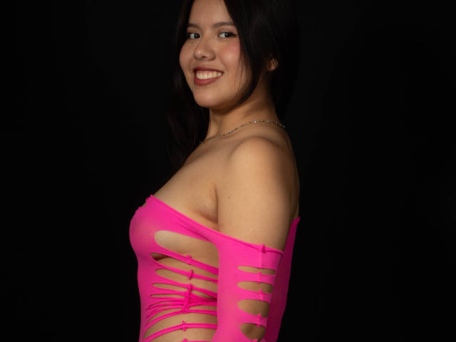MarielQueenX69 with dark hair, smiling, wearing a hot pink, cut-out top exposing her tits and side of her ass, against a black background.