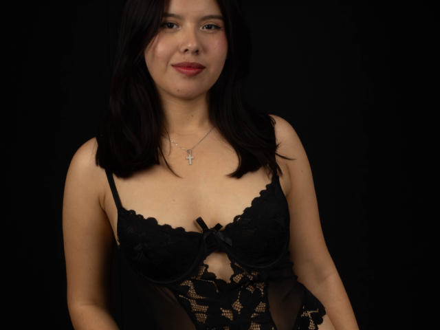 MarielQueenX69 with dark hair, wearing black lace lingerie with bow, showing small tits, cute face, slight smile, cross necklace, black background, soft lighting.