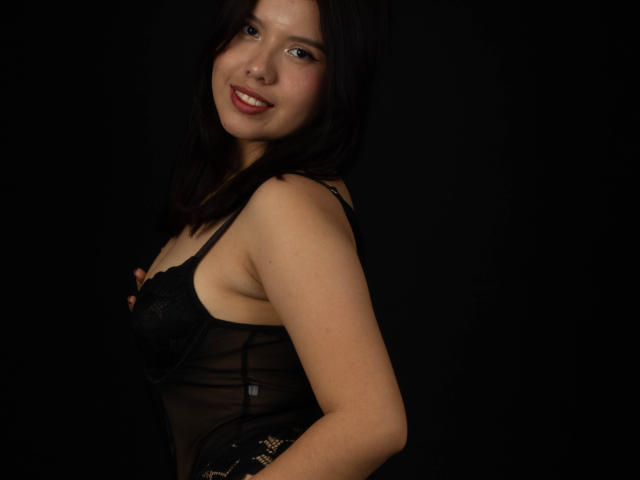MarielQueenX69 with dark hair, smiling, wearing a sheer black lingerie top that shows her tits, against a black background. She's looking over her shoulder.