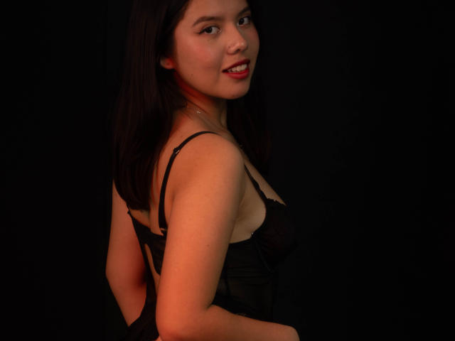 MarielQueenX69 with black lace lingerie, showing her tits, smiling at the camera, dark brown hair, red lipstick, black background, soft orange light.