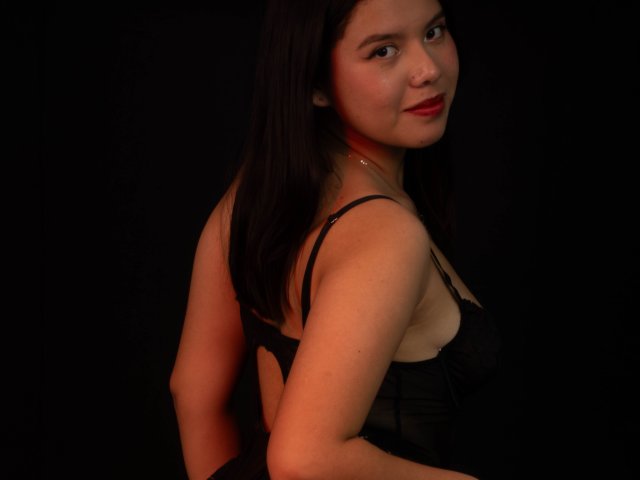 MarielQueenX69 with medium tits, black lingerie, and red lipstick, looking over shoulder, dark brown hair, slight smile, black background, soft lighting, confident, sexy, slightly curved back.