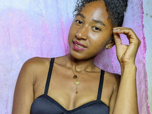 HotChrstine69, avec ses cheveux noirs bouclés, sa peau brune et ses petites seins, portant un soutien-gorge noir, un collier en or, et avec un fond rose, souriant tout en touchant sa tête avec sa main droite..