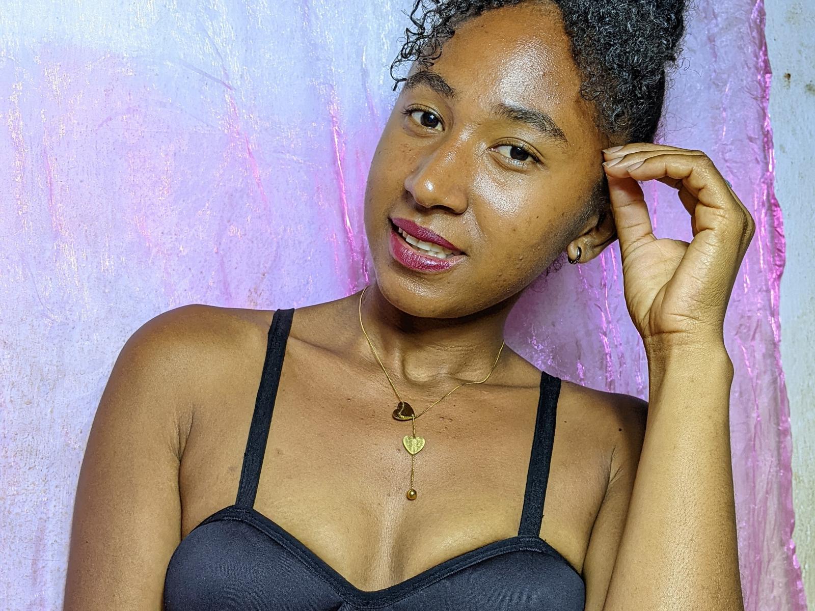 HotChrstine69 with curly black hair, brown skin, and small tits, wearing a black bra, gold necklace, and pinkish background, smiling with her right hand touching her head.