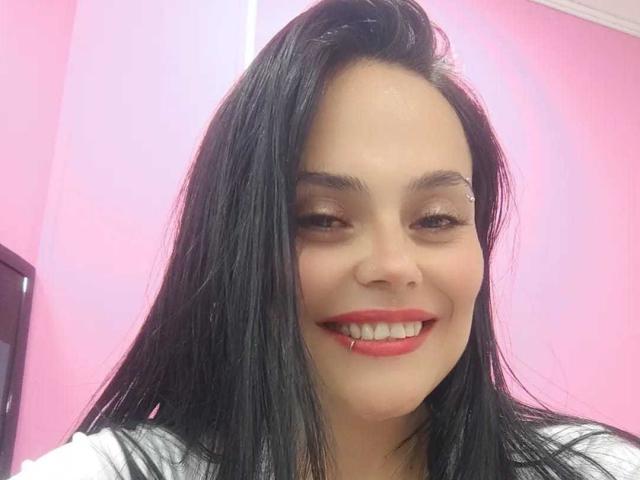 CharlotteCrofort with long black hair, smiling with red lipstick, white shirt, and visible teeth, has a cute face and a small nose, looking slightly to the side.