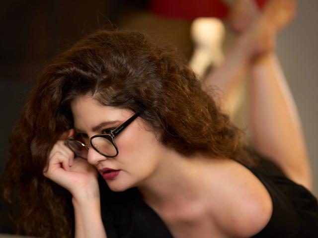 CelineBoyer with curly brown hair, black glasses, and red lipstick, holding glasses near her nose, wearing a black top, with her bare ass and legs in the blurred background.