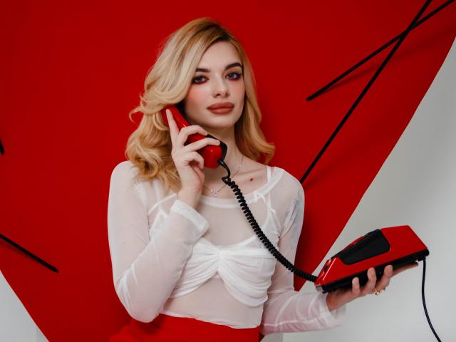 Adele Clifford mit blonden Haaren, die ein durchsichtiges weißes Oberteil trägt, hält ein rotes Telefon, spricht damit, roter Hintergrund, roter Rock, schwarzes Kabel, leichtes Lächeln, selbstbewusst..