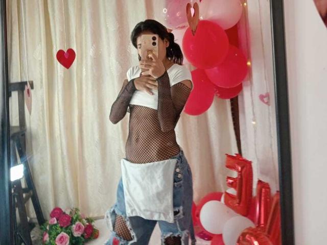 AnnieDarling with dark hair, wearing a fishnet top, white tee, and ripped jeans, standing in front of a mirror, holding a phone, surrounded by red balloons.