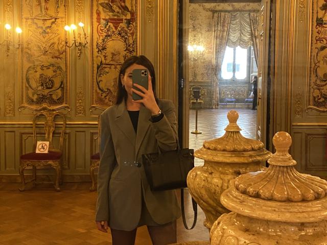 Dionisia with medium tits, wearing a grey blazer and black handbag, taking a mirror selfie in a fancy room, holding a green phone, standing near ornate marble jars.