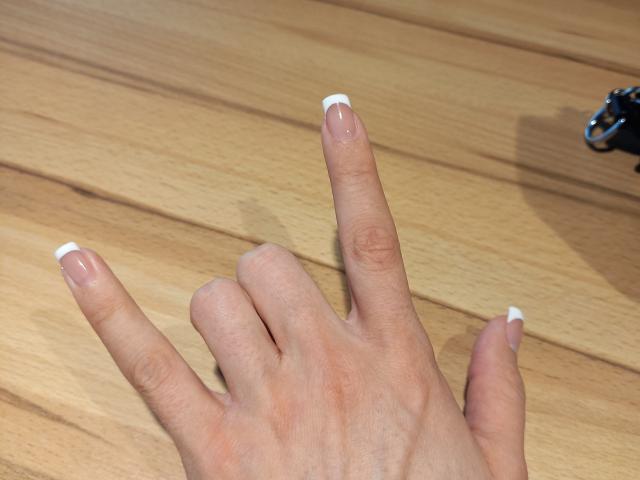 BellaJulls' hand with French-manicured nails, fingers spread, pointing upward. She has light skin, and the wood floor is visible in the background. Her pinky's slightly curled.