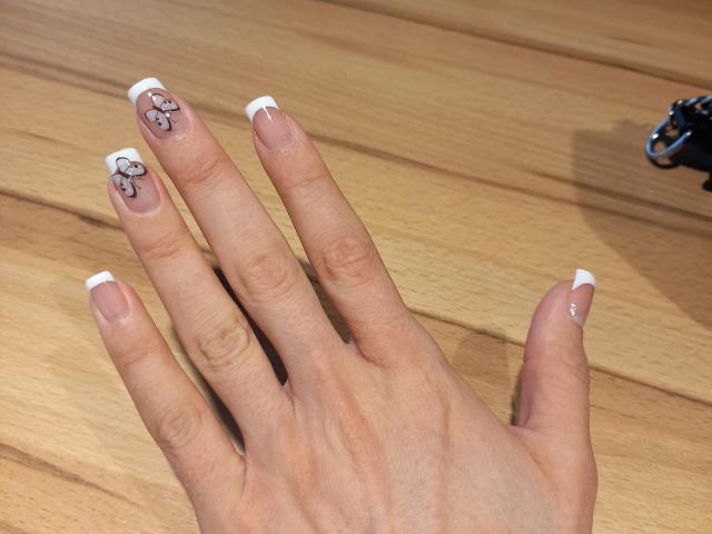 BellaJulls' hand with French tip nails, white tips, and cute cartoon cat tattoos on her index and middle fingers, sitting on a wooden floor.