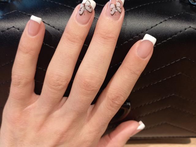 BellaJulls' hand with French tips, her nails have cute, small, glittery designs on the top, resting on a black, quilted leather surface. Her skin is light and smooth.