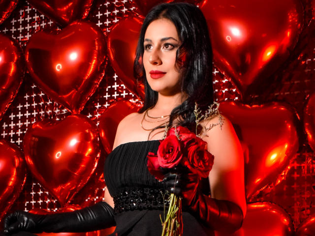 MaddySmit con un abito nero, rose rosse e guanti neri, che tiene fiori, palloncini a forma di cuore rossi e uno sfondo scintillante, sorridente, con capelli neri ondulati e un tatuaggio sulla spalla destra..