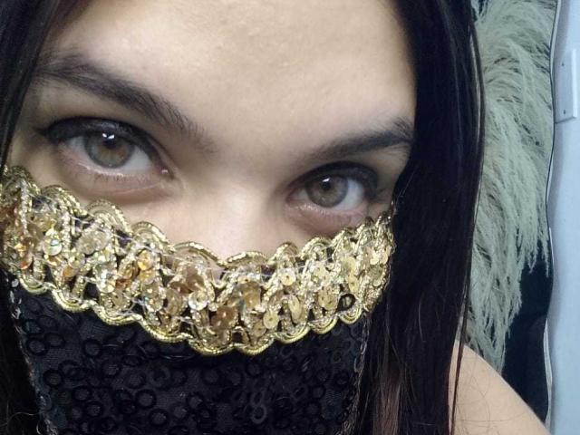 Les yeux d'AniisaAli, avec leurs pupilles brun foncé, sont encadrés par un masque en dentelle doré et noir qui recouvre son nez et sa bouche. Ses longs cheveux bruns sont visibles..