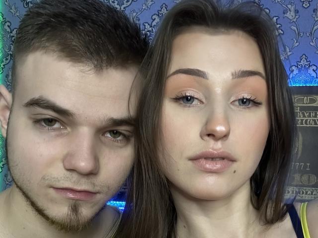 Vikki has glossy pink lips and a small beauty mark below her nose. George has short brown hair and a goatee. Vikki's eyes are lined with black eyeliner.