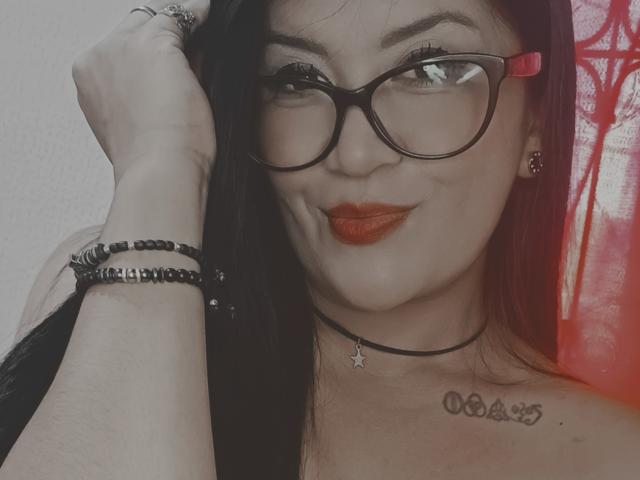 Daphne Rubine, com óculos pretos, cabelo ruivo, batom laranja, colar preto, tatuagem de estrela, pulseiras e uma mão no cabelo, sorrindo. Ela tem um rosto bonito e seios pequenos..
