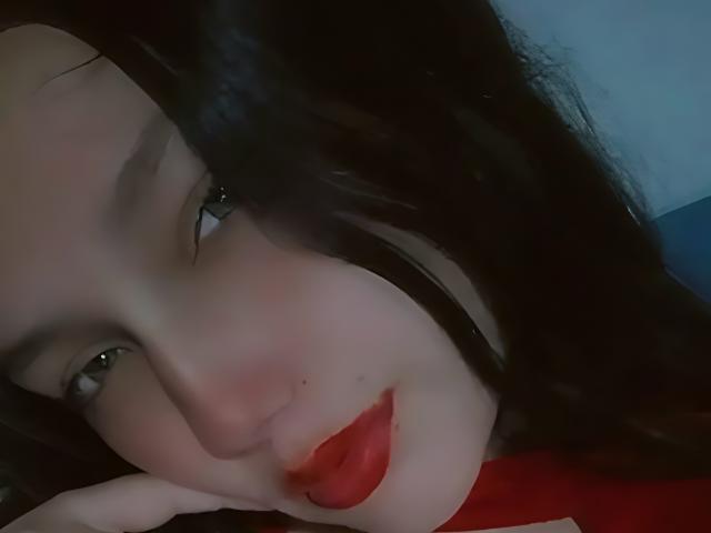 JenniferLw's face, dark wavy hair, pale skin, green eyes, red lipstick, slight blush, resting chin on hand, subtle smile, soft light, close-up, gentle expression, slightly tilted head.