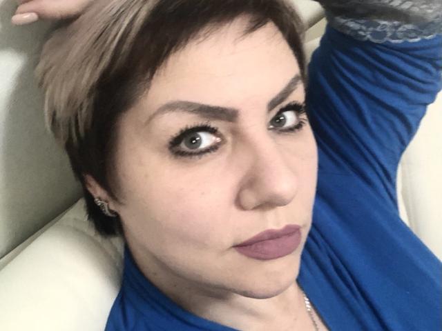 AngelinaGlam with short, brown-to-blonde hair, blue eyes, and purple lipstick, wearing a blue top, looking at the camera with a slight pout. Her hand's in her hair.