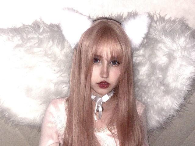 KaylaWow has long pink hair, white cat ears, fluffy white wings, and a white choker with a ribbon. She's wearing a pink, lace-trimmed top with red lipstick.