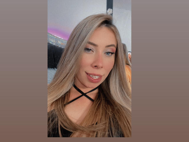 JennyLawry with long blonde hair, blue eyes, and pink eyeliner, smiling slightly, wearing a black strappy top, has cute tits, and a sexy face, in a selfie.