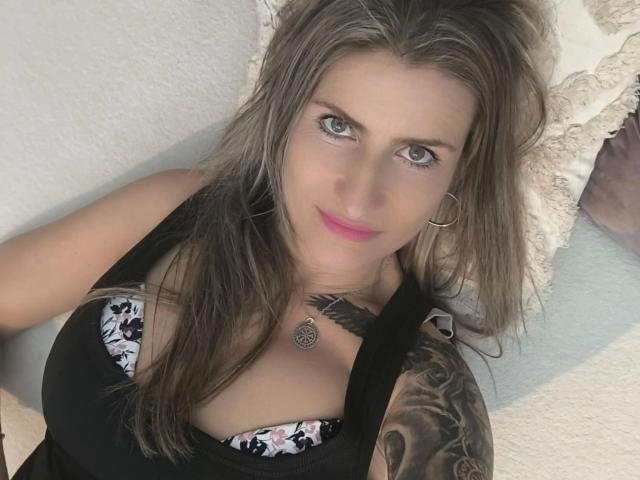 KatyStar-hot with blonde hair, green eyes, pink lipstick, black tank top, tattoo on right arm, and a tattoo necklace, looking up with a slight smile. Boobs visible in tank top.