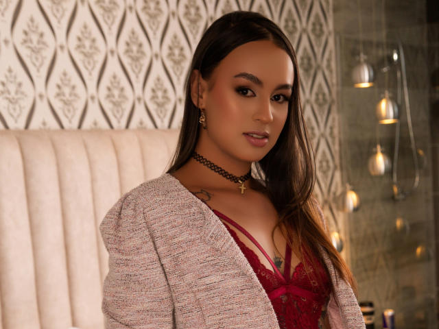 LaylaRays with brown hair, wearing a red lace bra and pink cardigan, choker necklace, earrings, and subtle makeup, showing small tattoos on her chest. Boobs visible through lace.