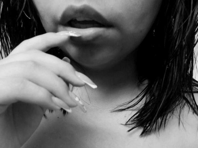 Lilys vollfotige Lippen sind leicht geöffnet, und ihre langen, gepflegten Finger der rechten Hand berühren sanft ihre Unterlippe. Sie hat feuchtes, dunkles Haar und eine zarte, helle Haut..