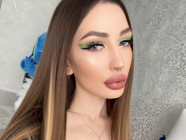 Martinik has long brown hair, green and black winged eyeliner, a nose ring, and pink lipstick. Her tits are not visible, just her face. She's looking at the camera.