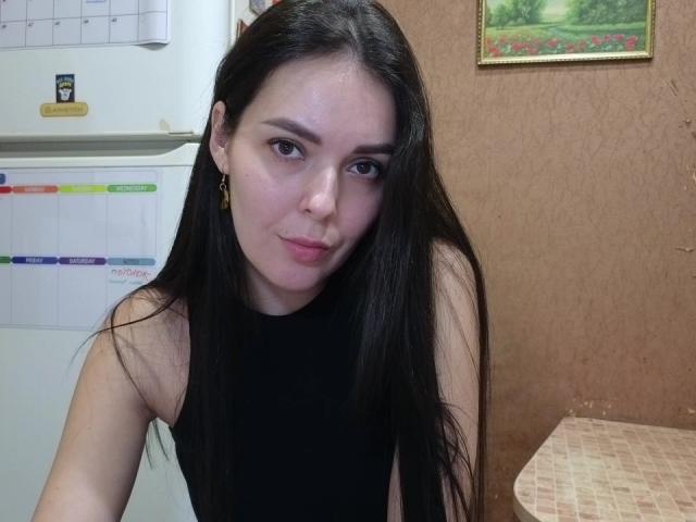 LissaBel with long black hair, wearing a black sleeveless top, looking at the camera with a slight pout, has small earrings, and is sitting near a fridge.