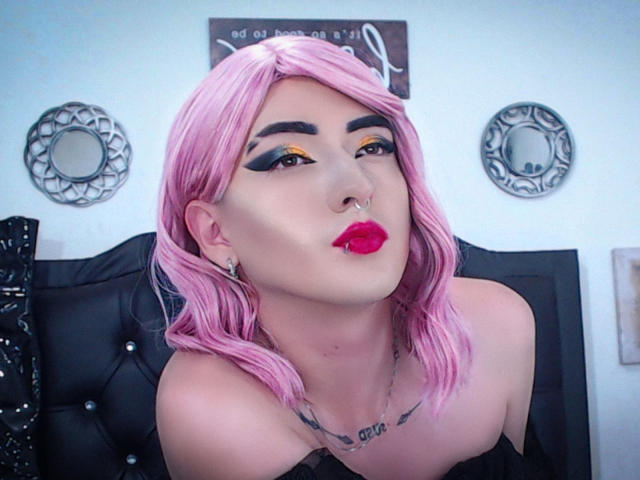 TokyoEvans, avec ses cheveux roses, son maquillage audacieux et son rouge à lèvres rouge, faisant la moue avec un anneau de nez, exhibant sa poitrine, un haut noir à épaules dénudées et un tatouage sur sa poitrine..