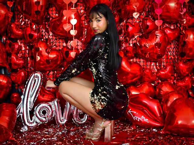 AriannaBrain est accroupie, vêtue d'une robe noire ornée de paillettes dorées, de talons rouges, entourée de ballons en forme de cœur rouges, avec de longs cheveux noirs et un sourire charmant..