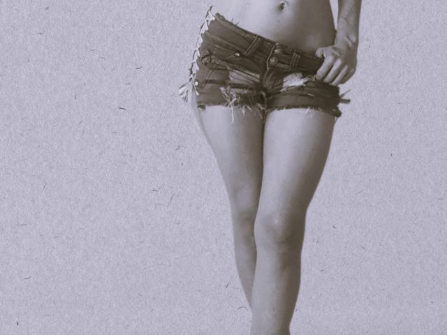 HarlyMia's ass is covered in frayed, dark denim shorts. Her hand is on her hip, showing off her slim, toned legs and flat stomach. Black-and-white photo.